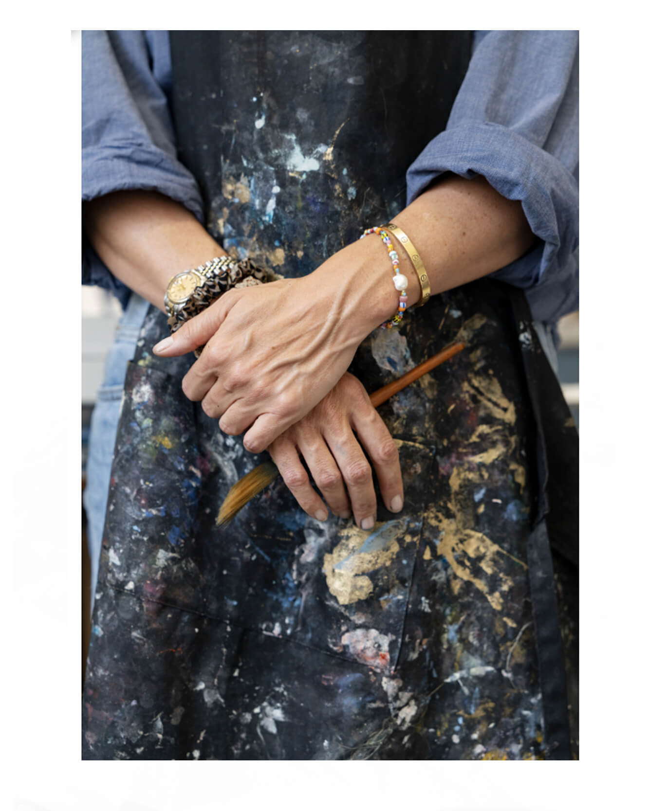 “Close-up of artist Caroline Tomlinson’s hands holding a paintbrush, wearing a paint-splattered apron in her London studio.”