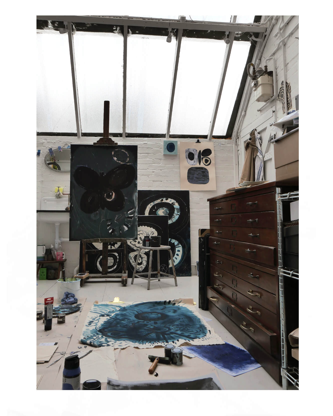 Artist Caroline Tomlinson’s London studio with paintings in progress, featuring abstract indigo works on canvas, art supplies, and natural light from a skylight.