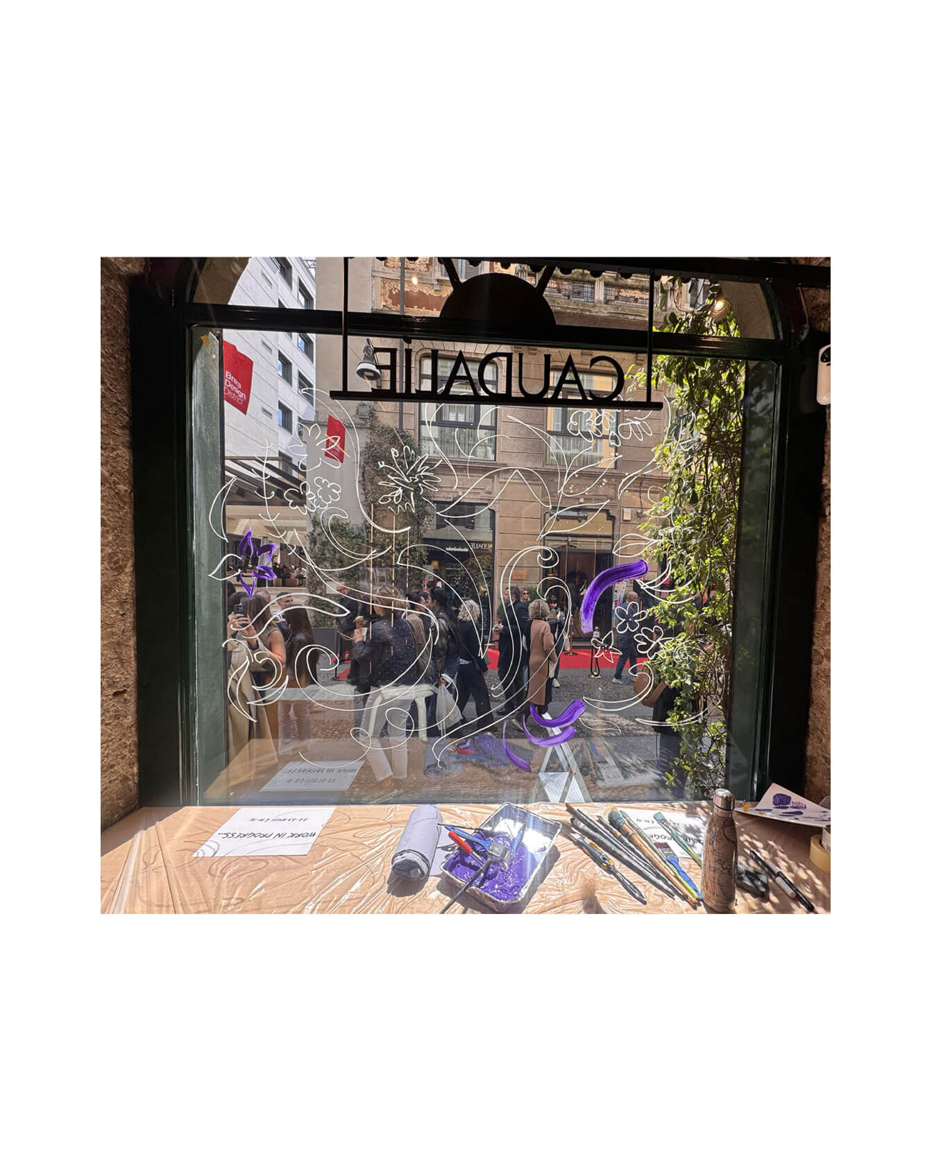 Artist live painting gestural botanical illustrations on large storefront windows during Milan Design Week for a luxury beauty brand, featuring fluid ink lines inspired by nature and fragrance.