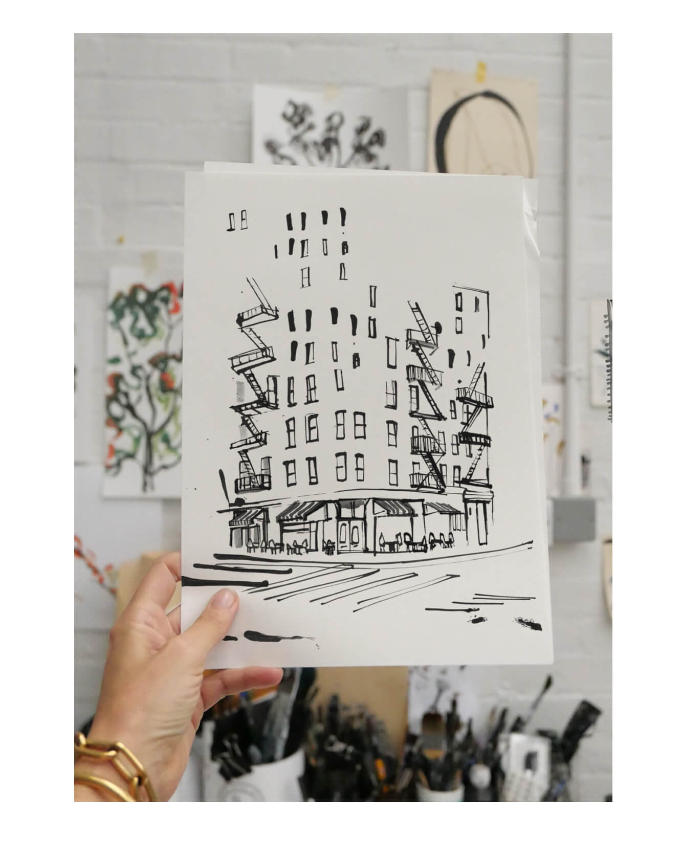 Hand-drawn ink illustration of a New York corner building with fire escapes and a street-level café, held up in the artist's studio.